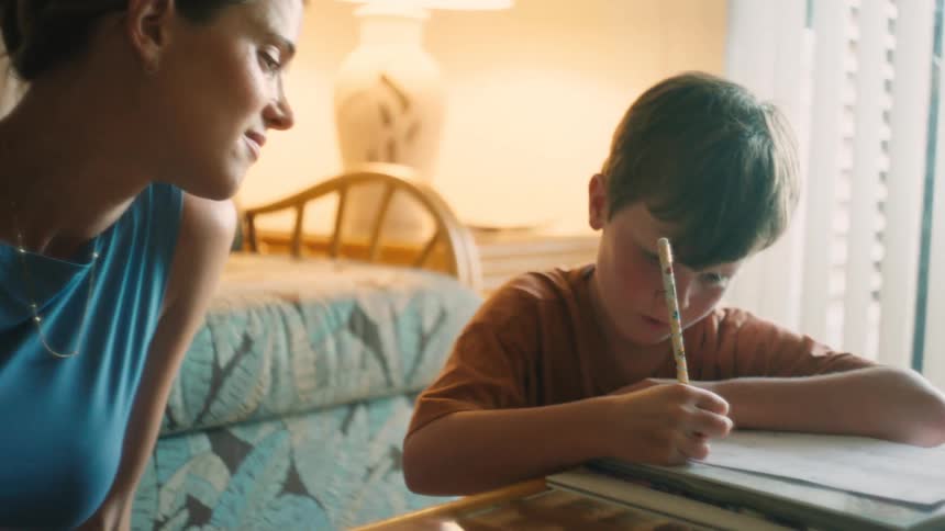 A caring mother smiles lovingly while watching her young son concentrate on his homework at a table in a cozy room