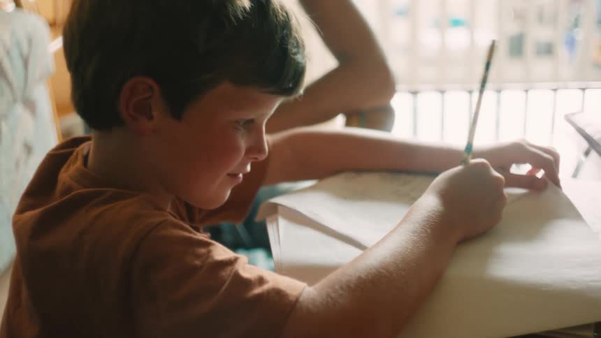 A happy young boy enjoys doing his homework, smiling as he writes with a pencil at a sunlit desk