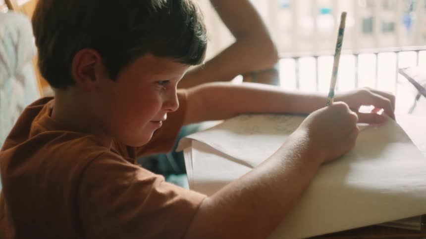 A happy young boy enjoys doing his homework, smiling as he writes with a pencil at a sunlit desk