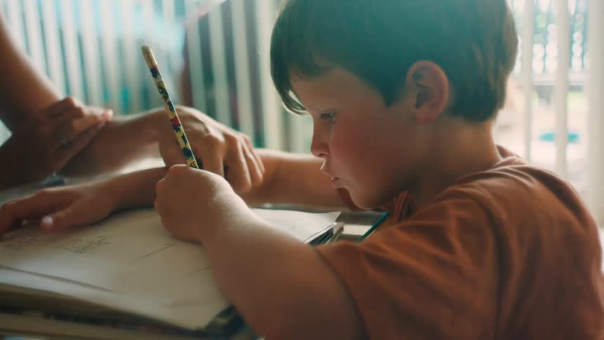 A focused young boy diligently works on his homework with a pencil as a parent offers guidance