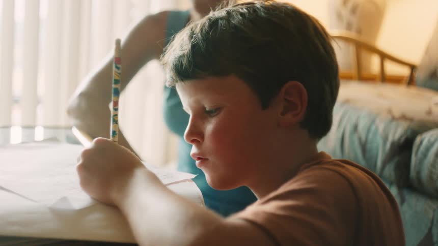 Boy concentrates on writing at a table in warm afternoon light while an adult helps nearby at home