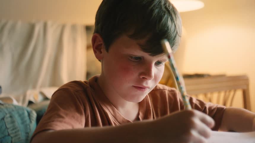 A focused young boy sits at a table and diligently works on his homework while a parent helps him