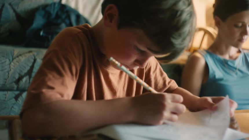 A focused young boy with freckles carefully writes on a paper with a pencil while doing his homework at home
