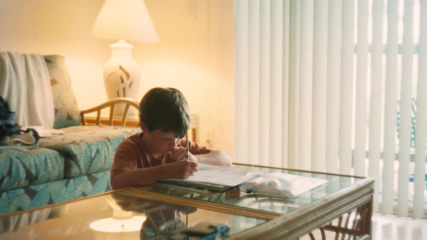 A caring mother sits with her young son at a coffee table, helping him with his homework in their cozy, sunlit living room