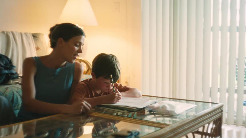 A caring mother sits with her young son at a coffee table, helping him with his homework in their cozy, sunlit living room