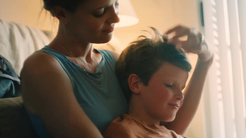 In a softly lit room, a mother lovingly caresses her son's hair and kisses his forehead in a heartwarming embrace
