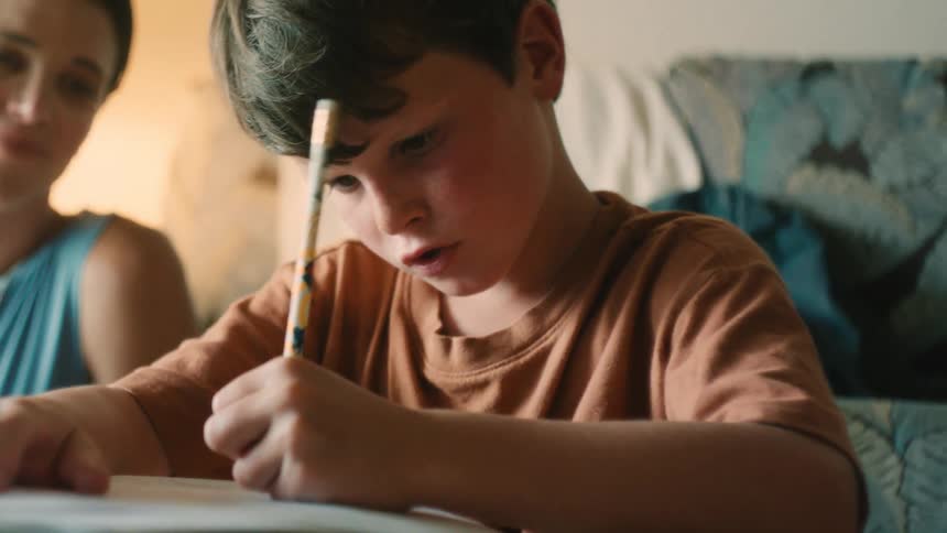 A young boy with freckles concentrates intently on his homework with a pencil to his forehead as his mother helps him