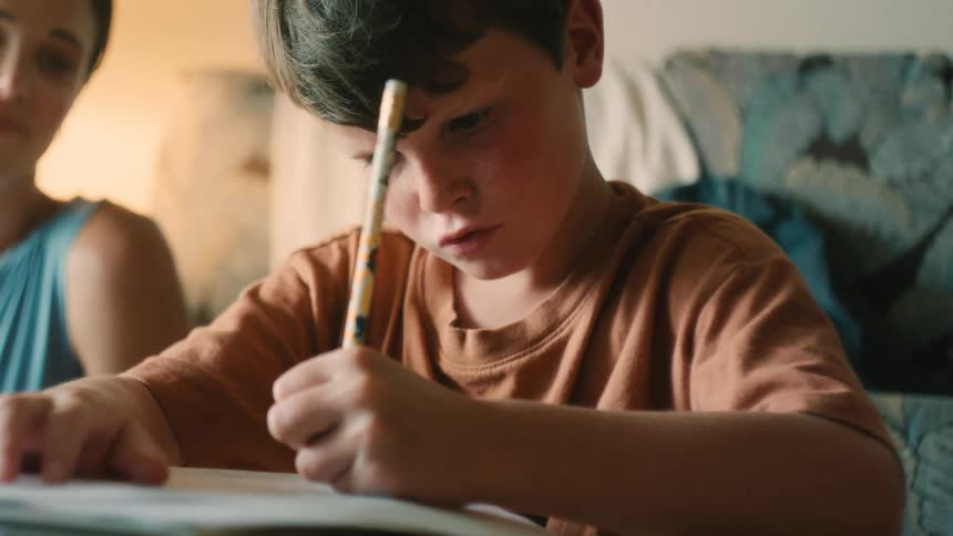 A young boy with freckles concentrates intently on his homework with a pencil to his forehead as his mother helps him