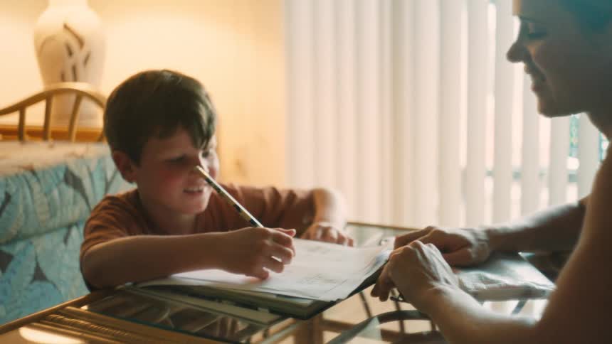 A mother smiles as she helps her playful son with his homework at a sunlit table in their home