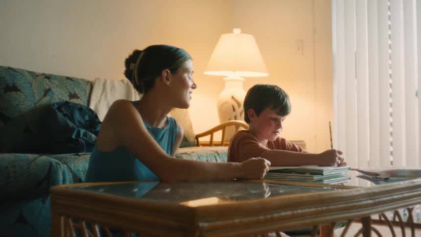 A caring mother smiles and encourages her young son as he works on his homework at the coffee table in their sunlit living room