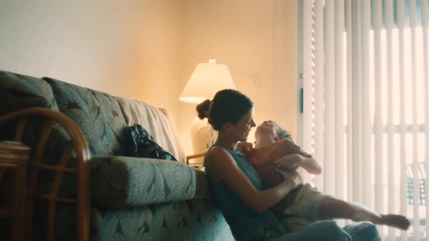 A mother playfully tickles her young son as they hug and laugh together on the floor of their sunlit living room