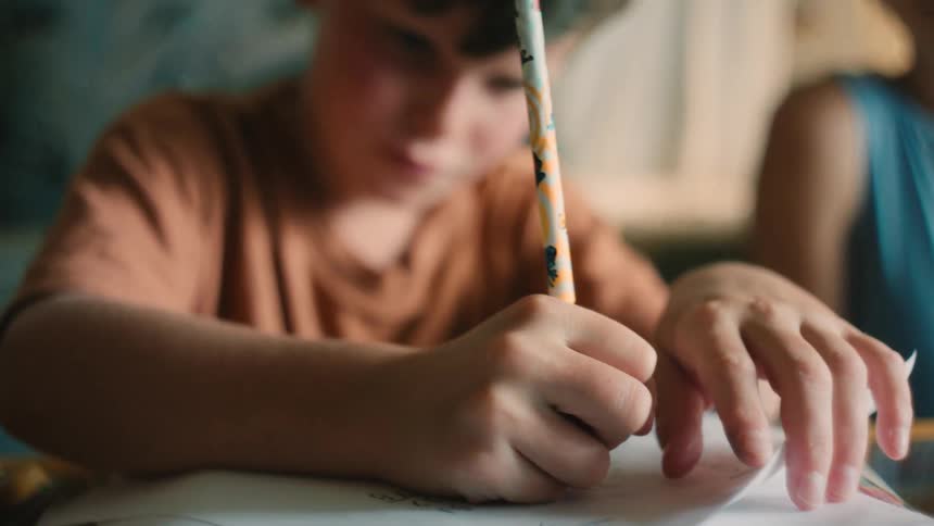 A young boy concentrates while writing with a pencil to complete his homework as his supportive mother watches over him