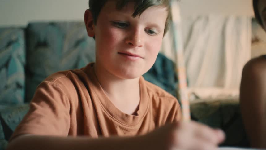 A young boy concentrates while writing with a pencil to complete his homework as his supportive mother watches over him