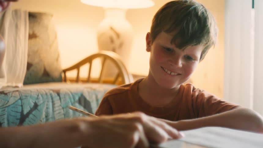 A mother helps her smiling son with his homework in a cozy, warmly lit room