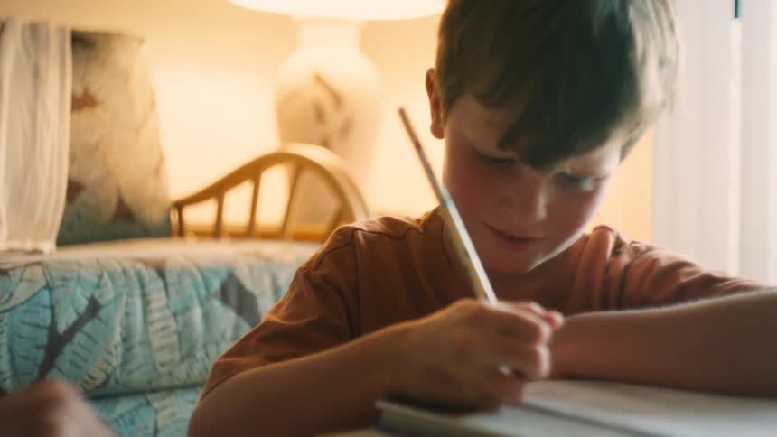 A mother helps her smiling son with his homework in a cozy, warmly lit room