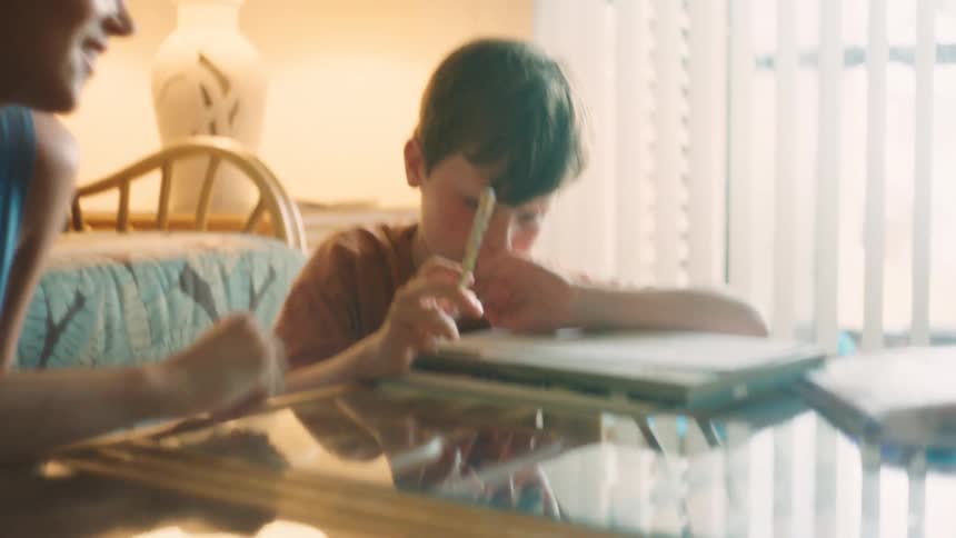A smiling mother patiently helps her young son with homework at a table in their warm, sunlit living room