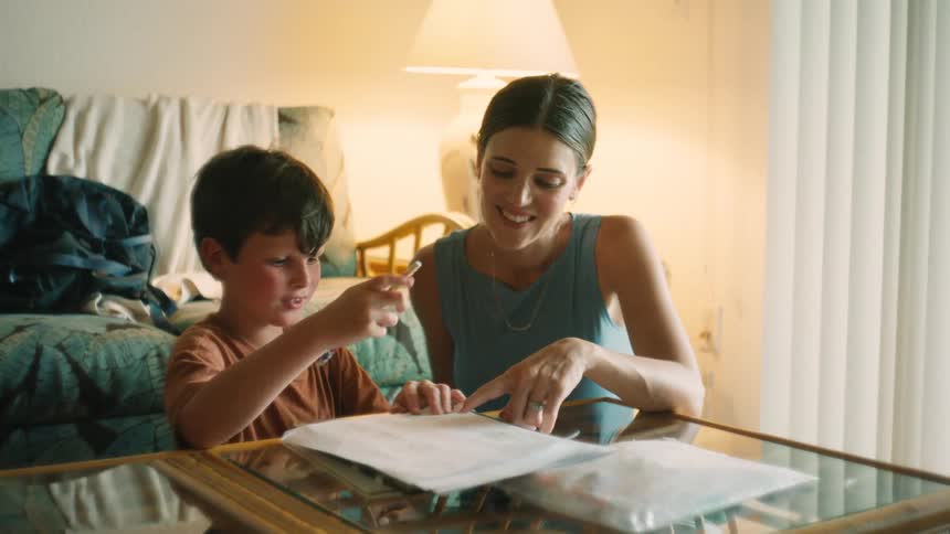 A loving mother helps her young son with his homework, smiling as he writes at the living room table
