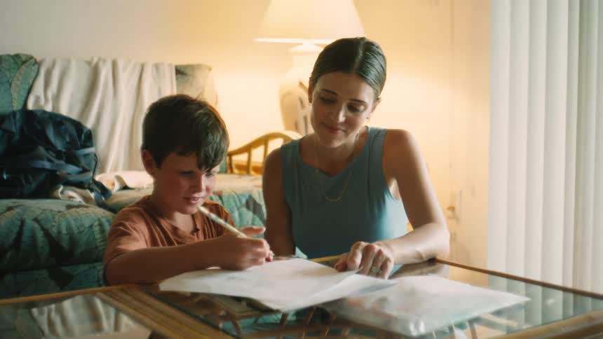 A loving mother helps her young son with his homework, smiling as he writes at the living room table