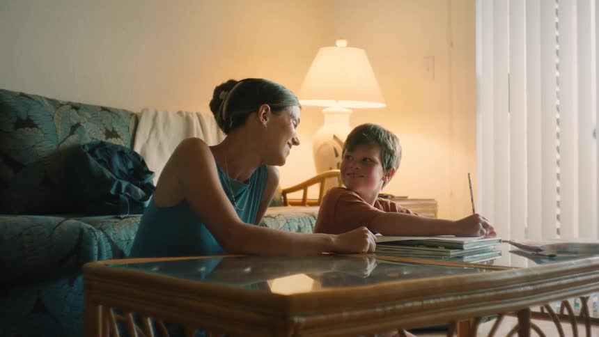 A proud mother lovingly watches her young son as he draws in a sketchbook at their cozy living room coffee table