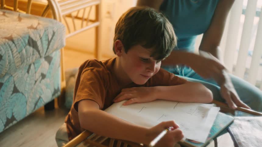 A caring mother kneels down to help her young son with his homework in their cozy, sunlit home