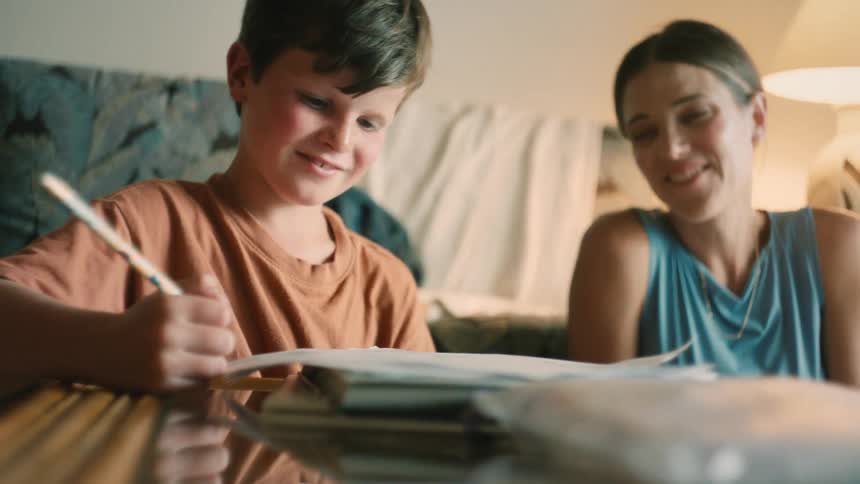 A caring mother proudly watches her young son as he concentrates on writing his homework at the table