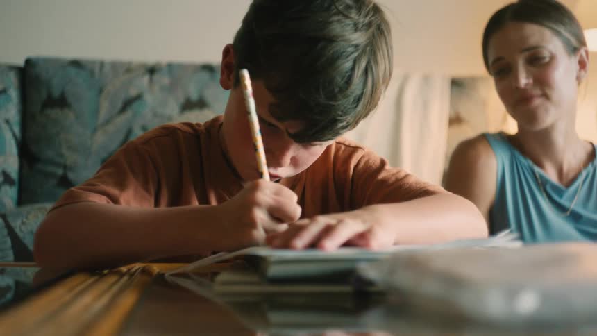 A caring mother proudly watches her young son as he concentrates on writing his homework at the table