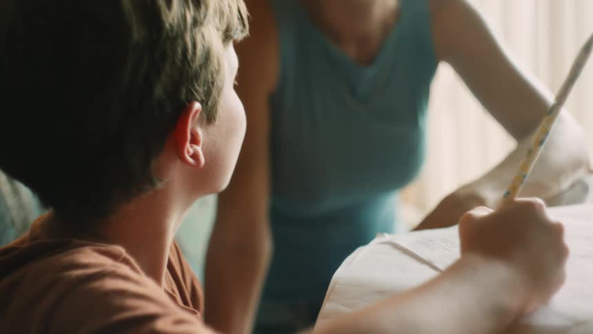 A young boy concentrates on his homework as his supportive mother sits beside him and points to his paper to help him