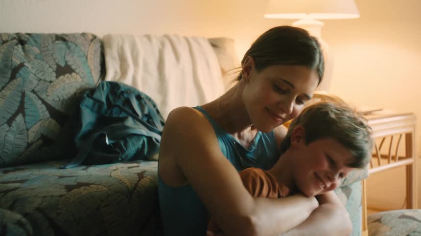 A mother tenderly comforts her smiling son, who becomes sad and begins to cry in her arms in a softly lit room