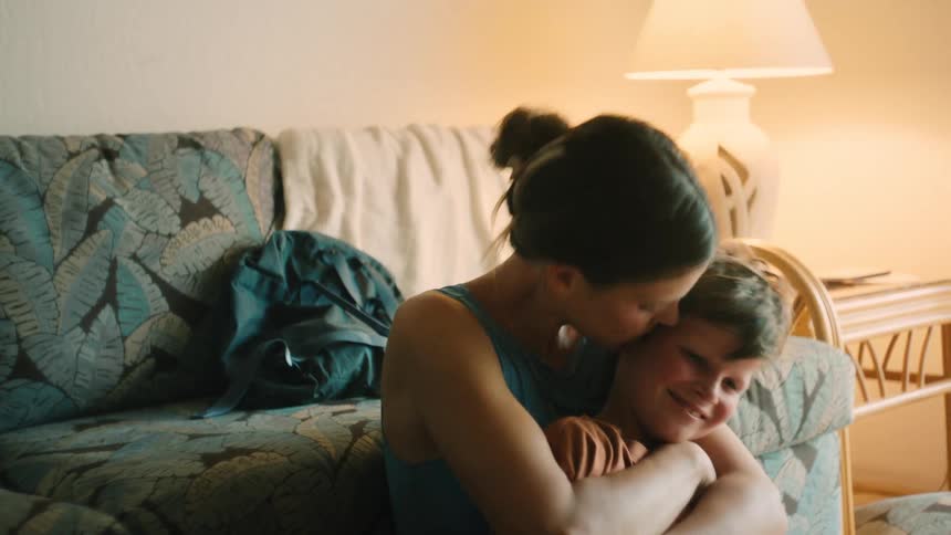 A mother tenderly comforts her smiling son, who becomes sad and begins to cry in her arms in a softly lit room