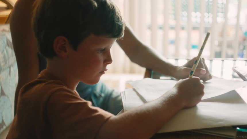 A mother patiently helps her young son with his schoolwork at a sunlit table in their home