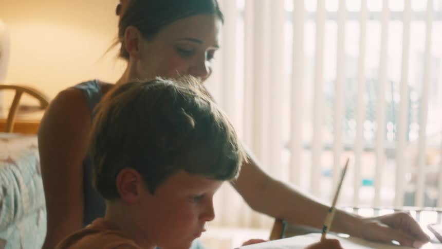 A mother patiently helps her young son with his schoolwork at a sunlit table in their home