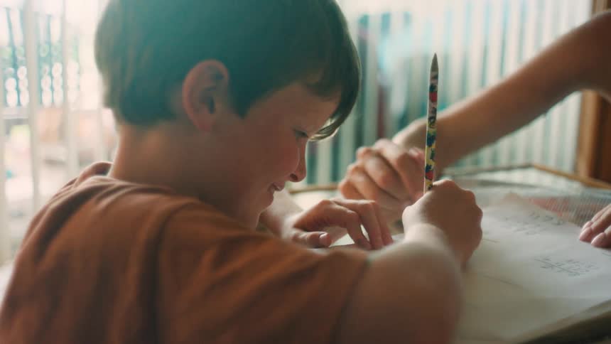 A young boy smiles while doing homework with his mother's help at a sunlit table, then refocuses with concentration