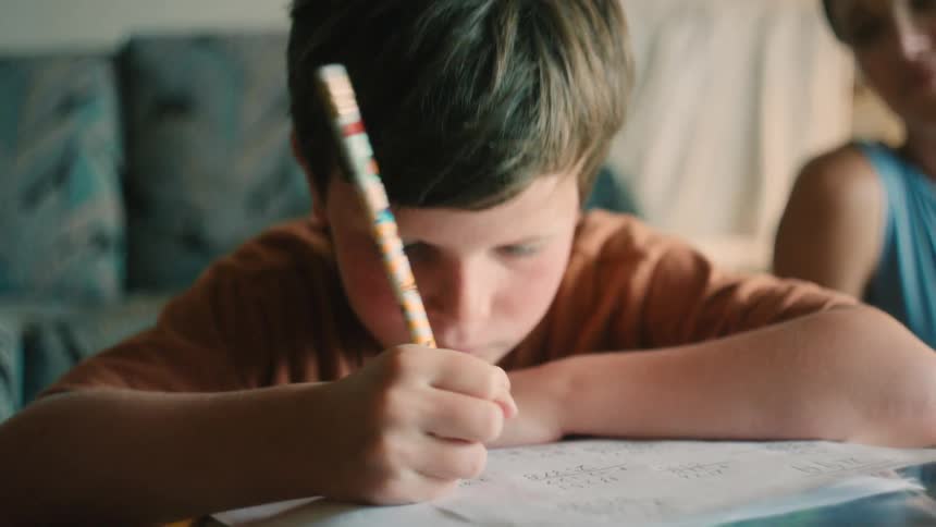 A young boy concentrates on his homework, carefully writing with a pencil as his mother watches