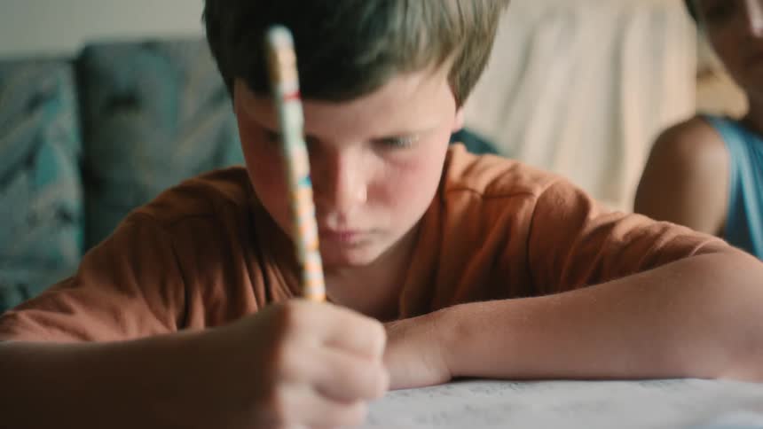 A young boy concentrates on his homework, carefully writing with a pencil as his mother watches
