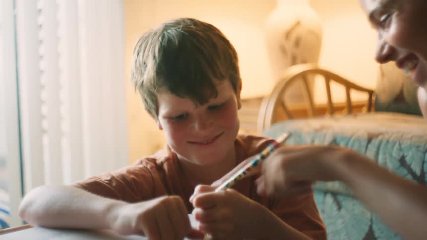 A close-up shot of a young boy smiling as his mother helps him with his homework in a softly lit room