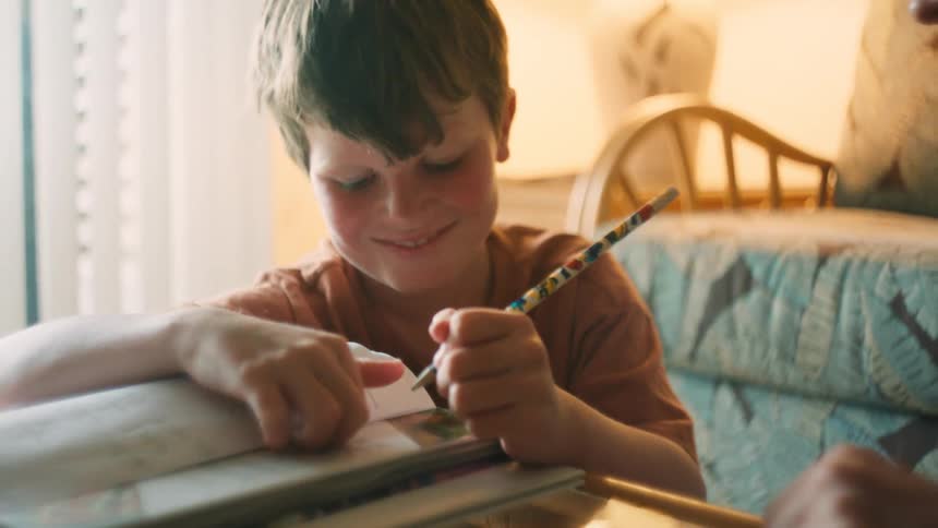 A close-up shot of a young boy smiling as his mother helps him with his homework in a softly lit room