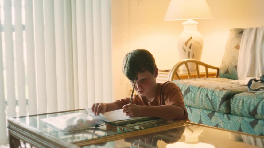 A young boy sits at a coffee table in a warmly lit living room, diligently writing or drawing on a piece of paper