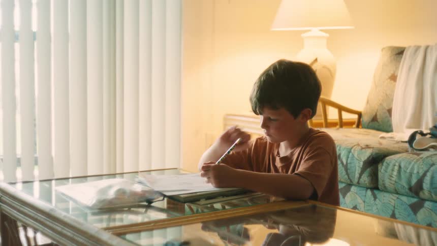 A young boy sits at a coffee table in a warmly lit living room, diligently writing or drawing on a piece of paper