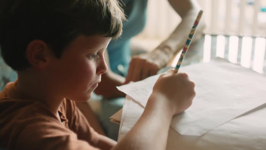 A young boy with freckles focuses intently on his homework, writing with a pencil as his mother helps him at the table in slow motion