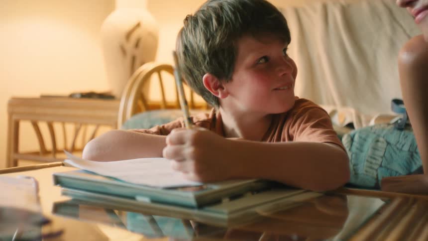 A smiling mother helps her young son with his drawing or homework, sharing a happy and playful moment together in a warmly lit room