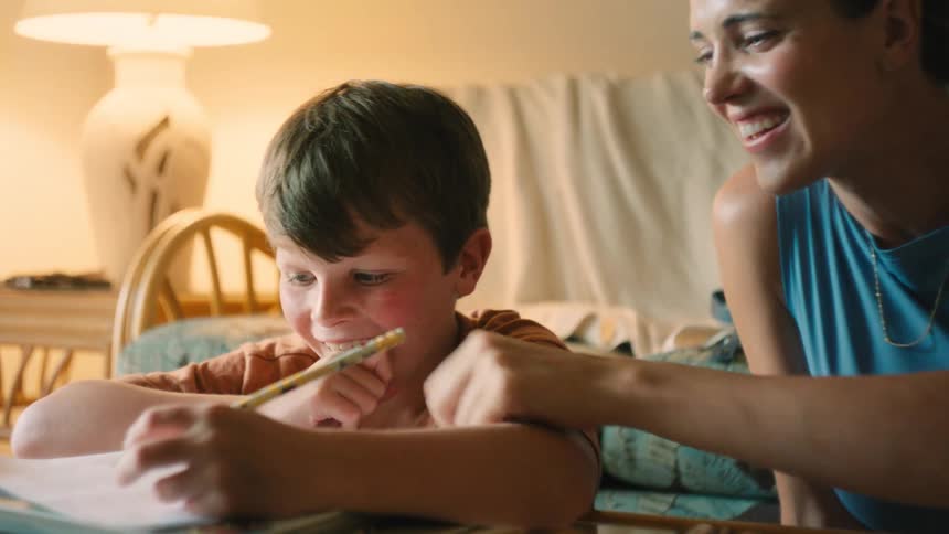 A smiling mother helps her young son with his drawing or homework, sharing a happy and playful moment together in a warmly lit room