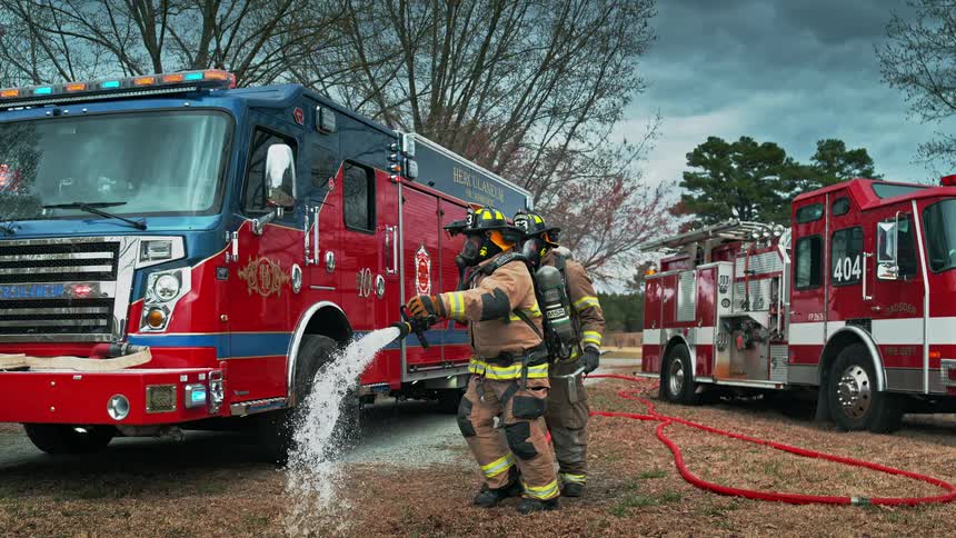 Two firefighters in full gear spray a powerful stream of water from a fire hose in dramatic slow motion with fire engines in the background