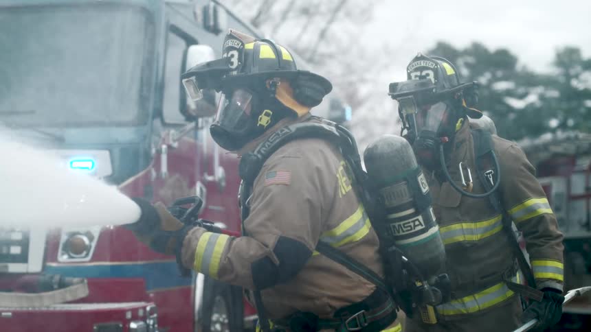 A firefighter in full gear aims a powerful fire hose, spraying water in dramatic slow motion with a fire truck in the background