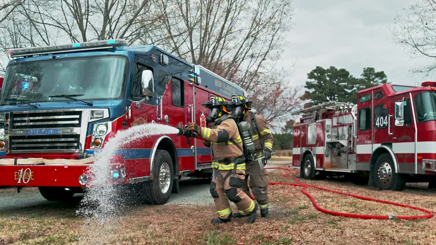 In a powerful slow-motion display, two firefighters brace themselves as they unleash a torrent of water from a fire hose with fire trucks in the background