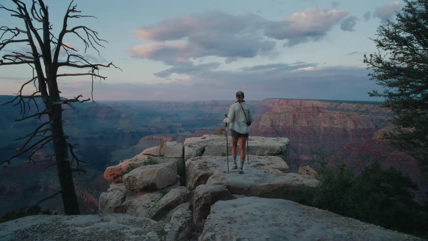 A female hiker with trekking poles walks to a rocky viewpoint to admire the majestic Grand Canyon at sunset