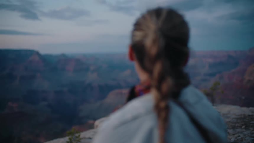A woman with her hair in a braided ponytail thoughtfully looks out over the Grand Canyon at dusk as the camera focus shifts to the vast landscape