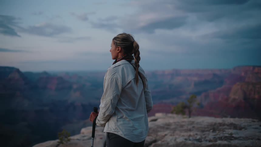 A female hiker with a trekking pole walks to a cliff's edge to admire the majestic view of the Grand Canyon at dusk
