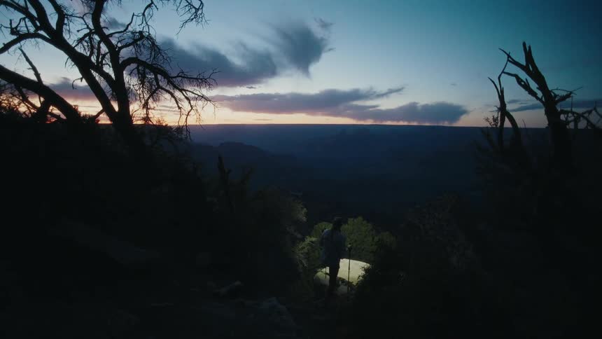 A hiker with a headlamp and trekking poles carefully descends a rugged trail at twilight, overlooking a vast canyon landscape