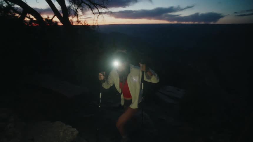 A determined woman with a headlamp and trekking poles hikes up a dark mountain trail in slow motion as the sun begins to rise