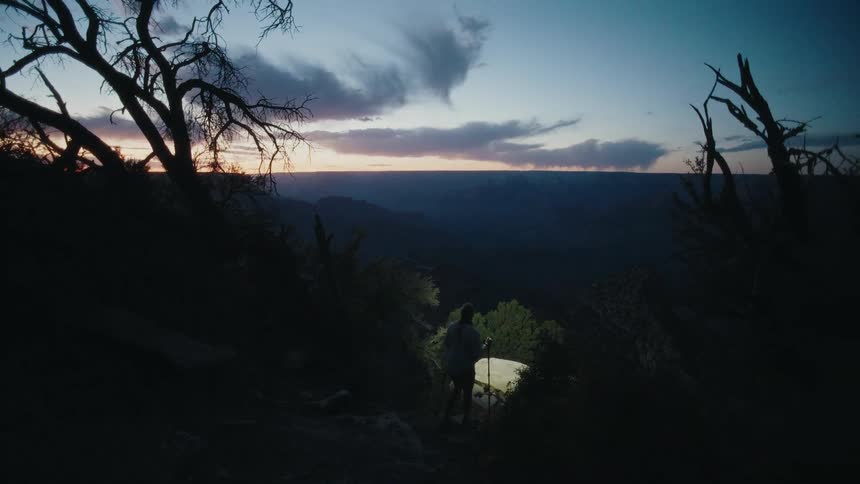 A solo female hiker with a headlamp navigates a dark trail down a mountainside at twilight to a scenic canyon overlook